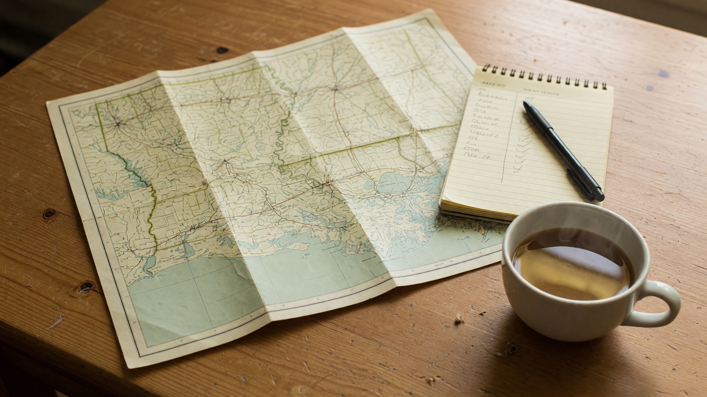 A folded Louisiana map, a two-column notepad, pen, and coffee on a wooden table
